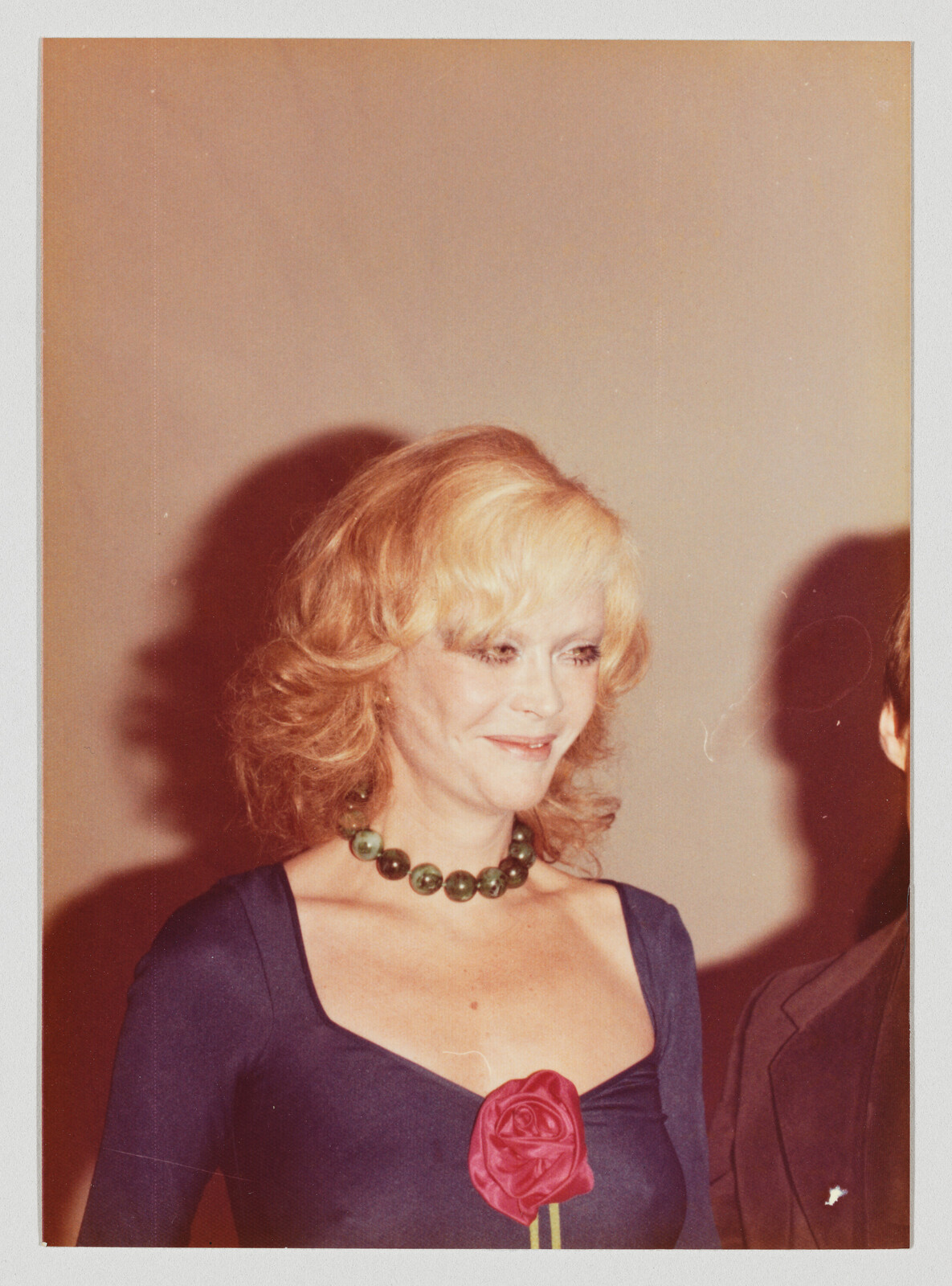 Blonde woman wearing a green beaded necklace and blue dress holding a red rose pinned to her chest.