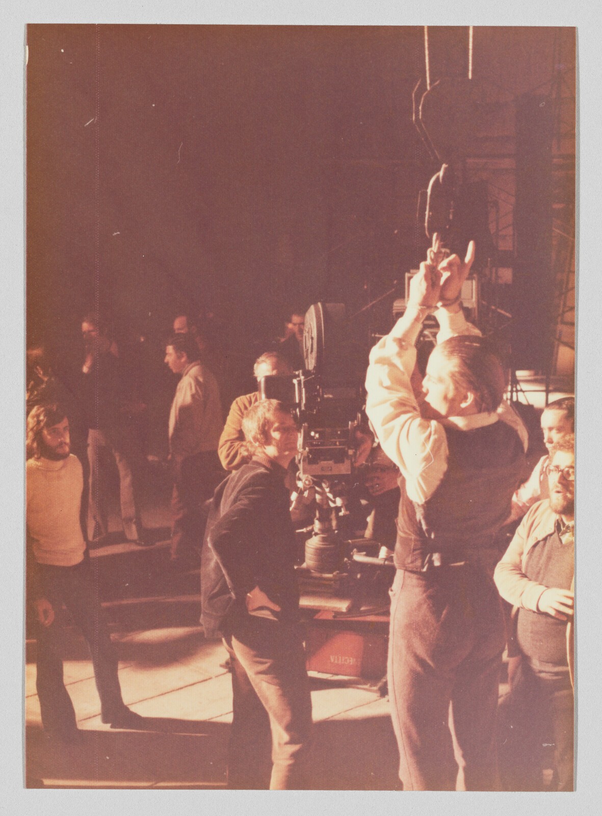 A film crew adjusts a large movie camera while a director raises his hands to signal.