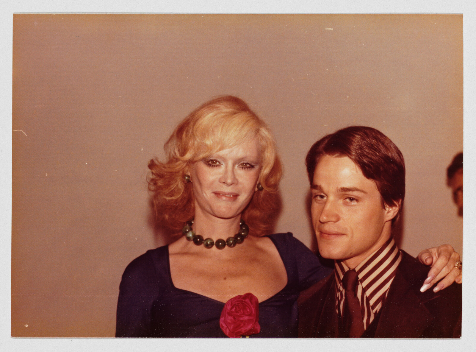 A woman wearing a green bead necklace and a rose holds her arm around a young man's shoulder.