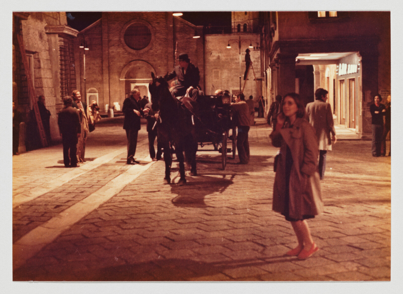 A woman in a coat stands on a dim street while a horse-drawn carriage and people gather nearby.
