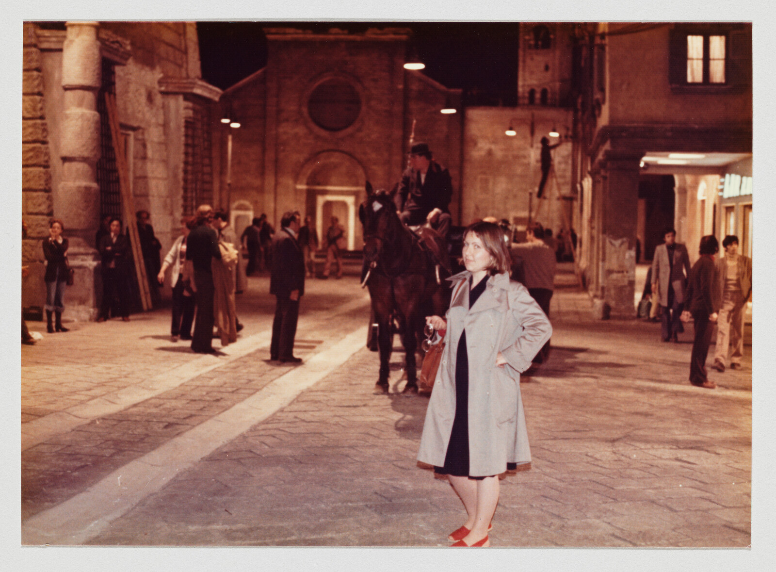 A woman in a trench coat stands hands-on-hip on a nighttime cobblestone street.
