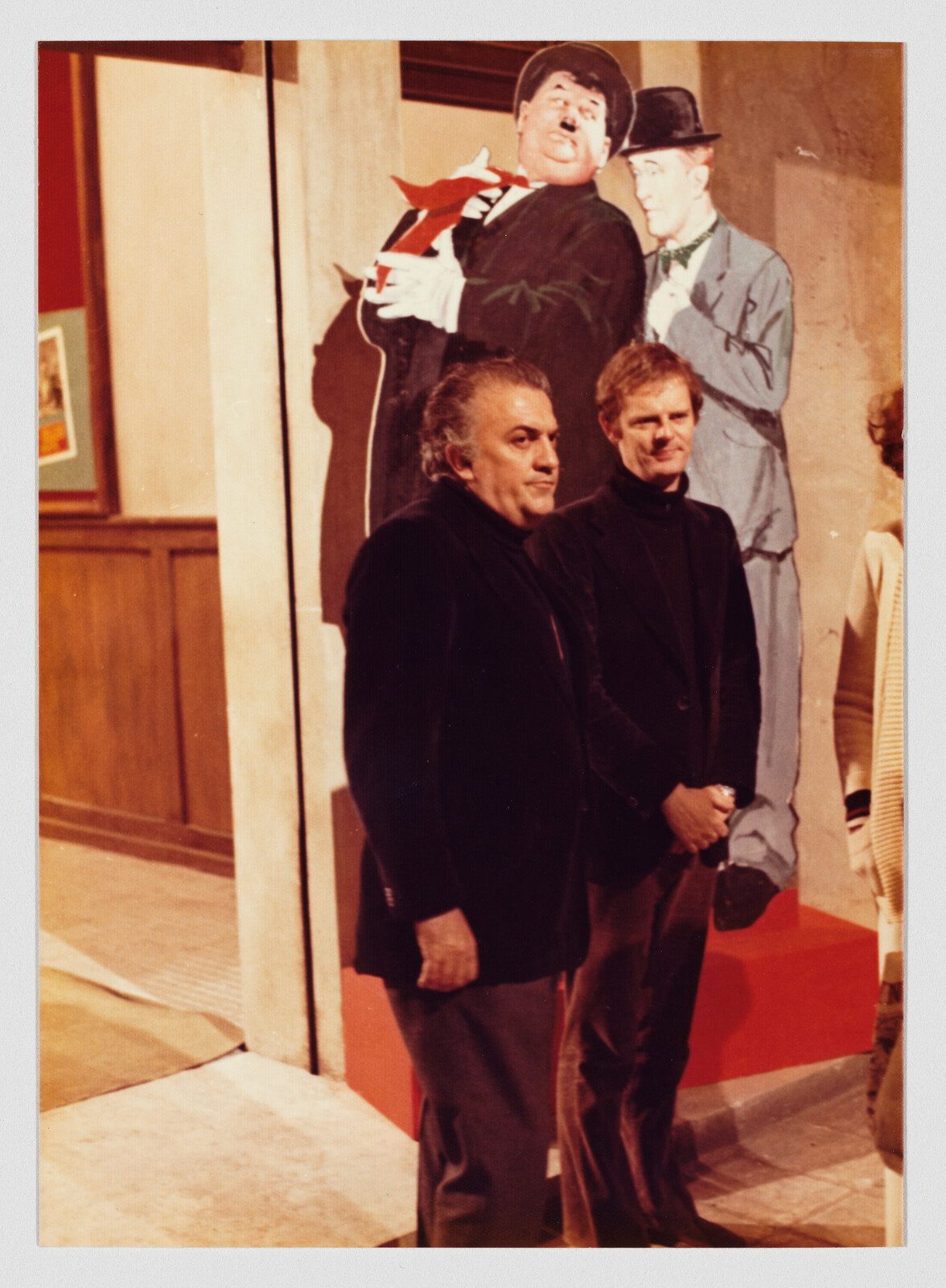 Two men in dark jackets stand side by side in front of large cartoon character cutouts.