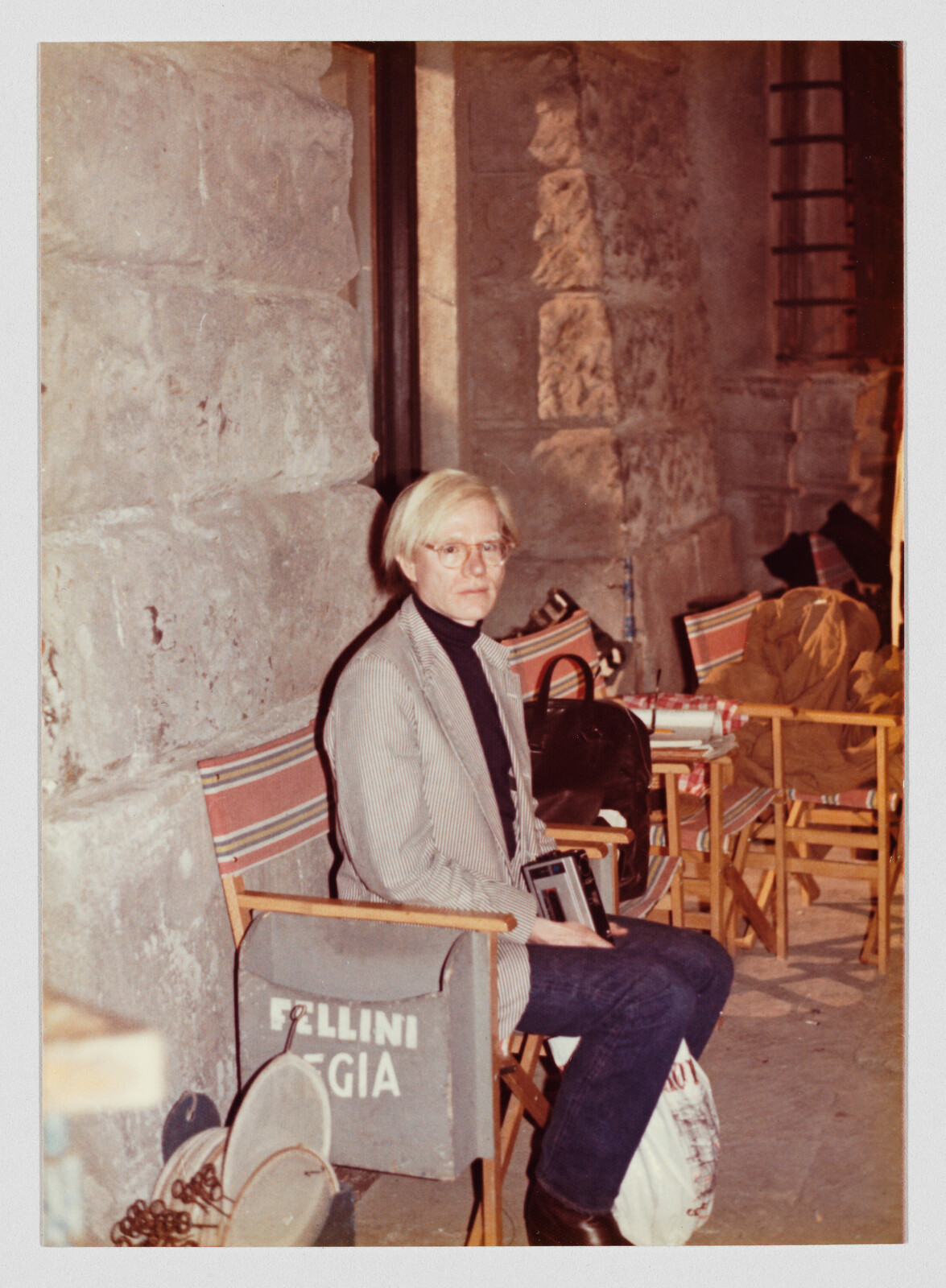 A person with light hair and glasses sits on a striped chair next to a chair labeled "FELLINI REGIA."