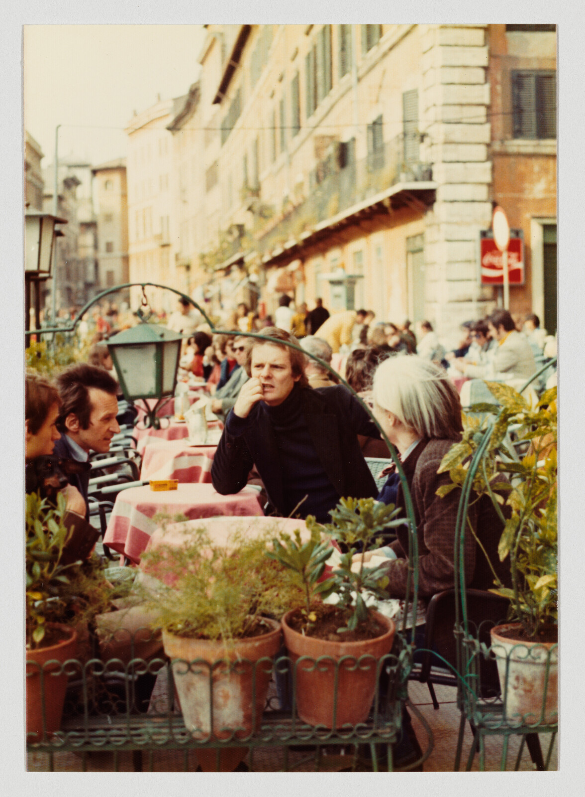 Man sitting at a crowded outdoor café table with others, resting hand on his chin.
