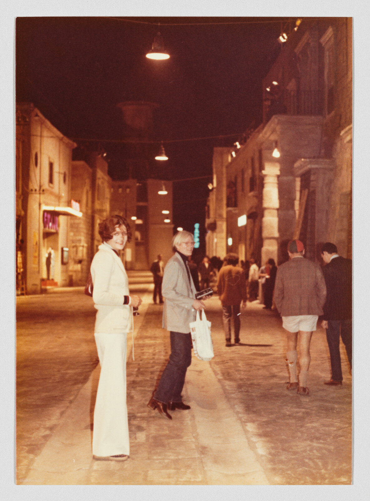A woman in white and a man pause and look back while walking down a dimly lit street.