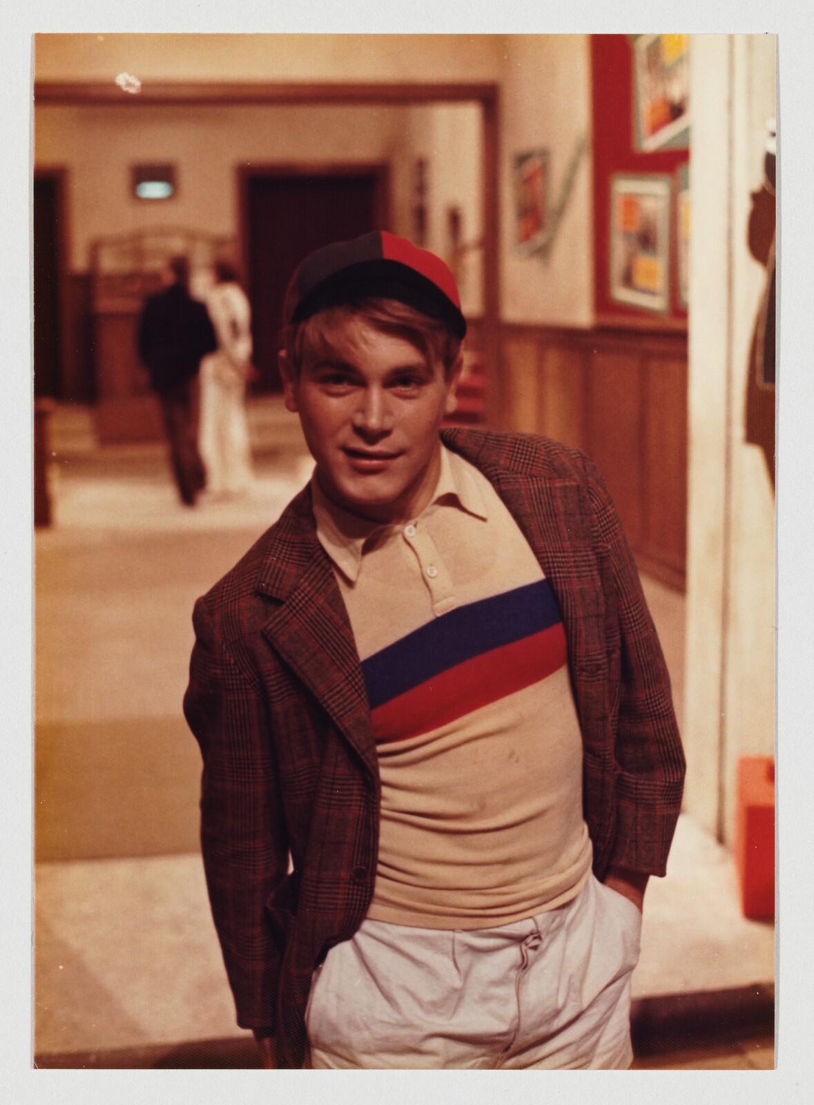 A young man wearing a striped shirt and cap leans forward in a hallway with hands in pockets.