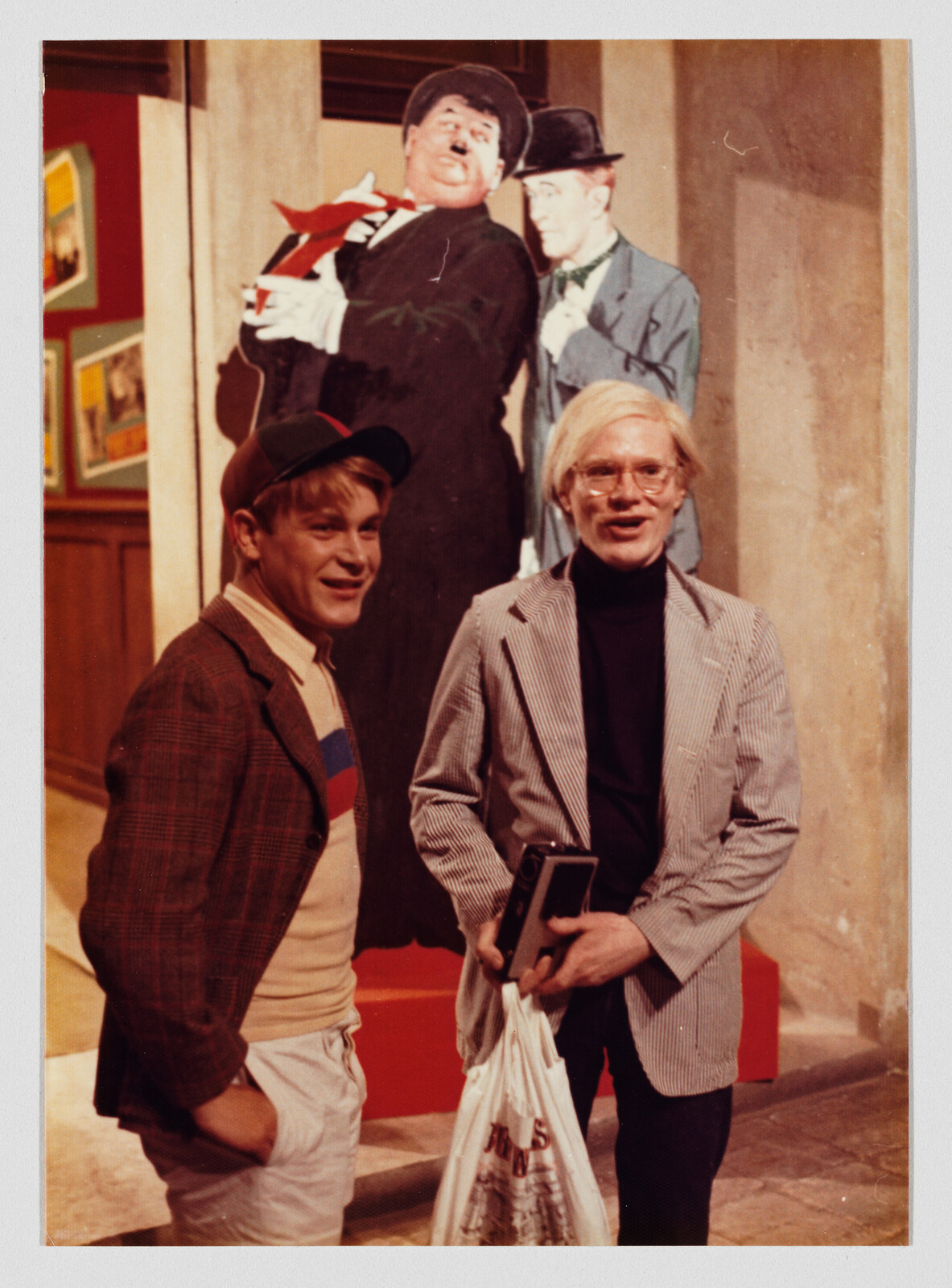 Two young men stand smiling by a museum exhibit, one holding a plastic bag and a book.