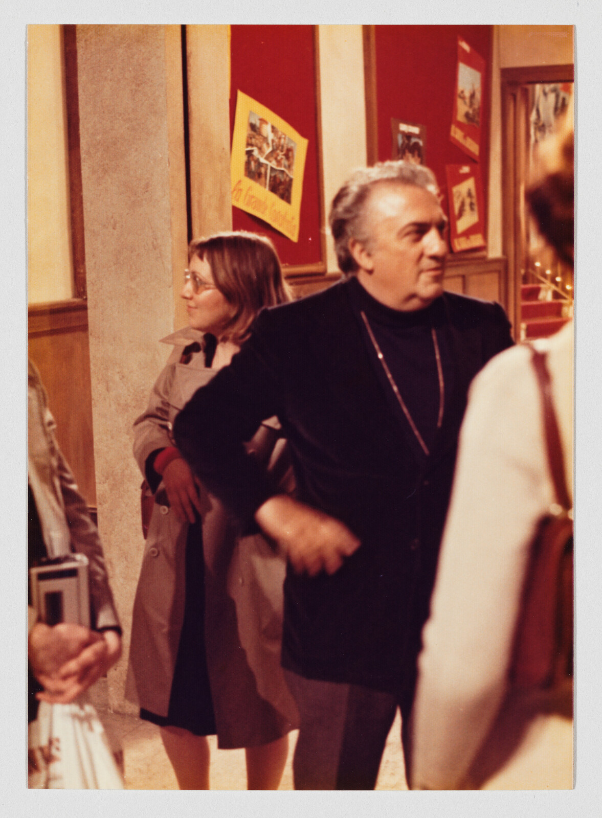 Older man in a dark jacket stands talking to others in a hallway with red posters.