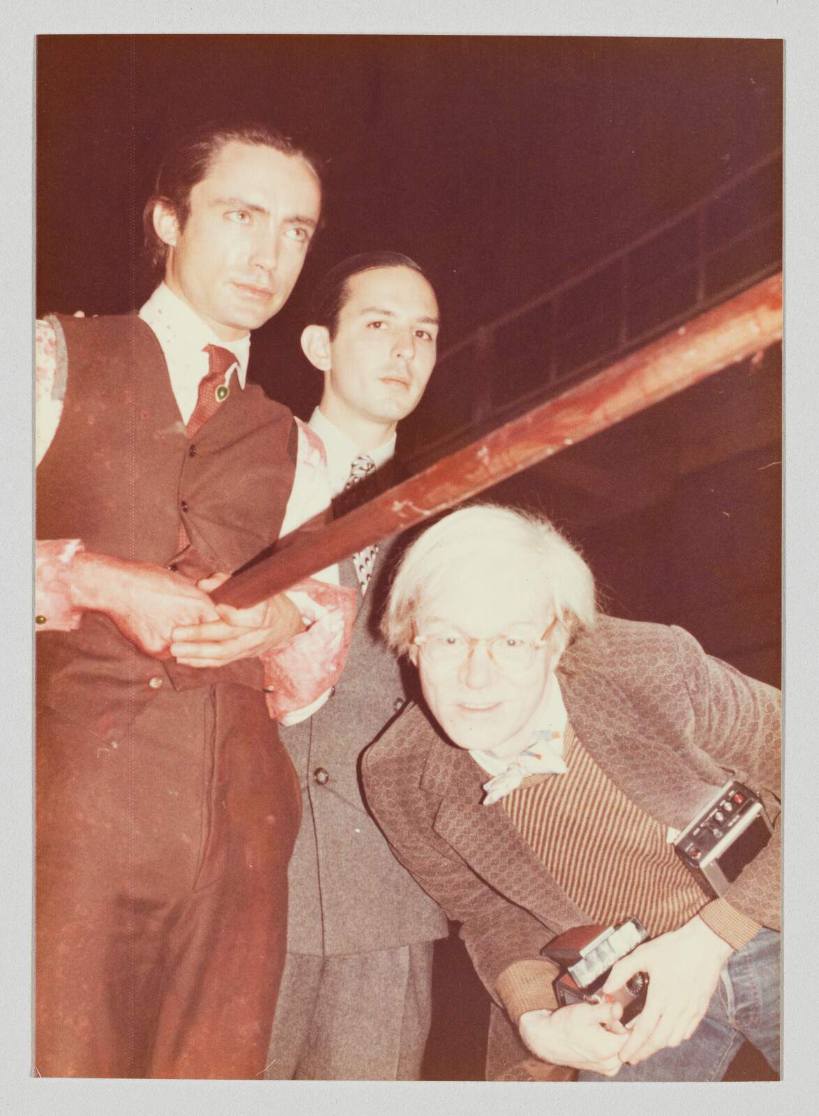 Three men lean toward the camera, with one crouching and holding a camera with a flash.