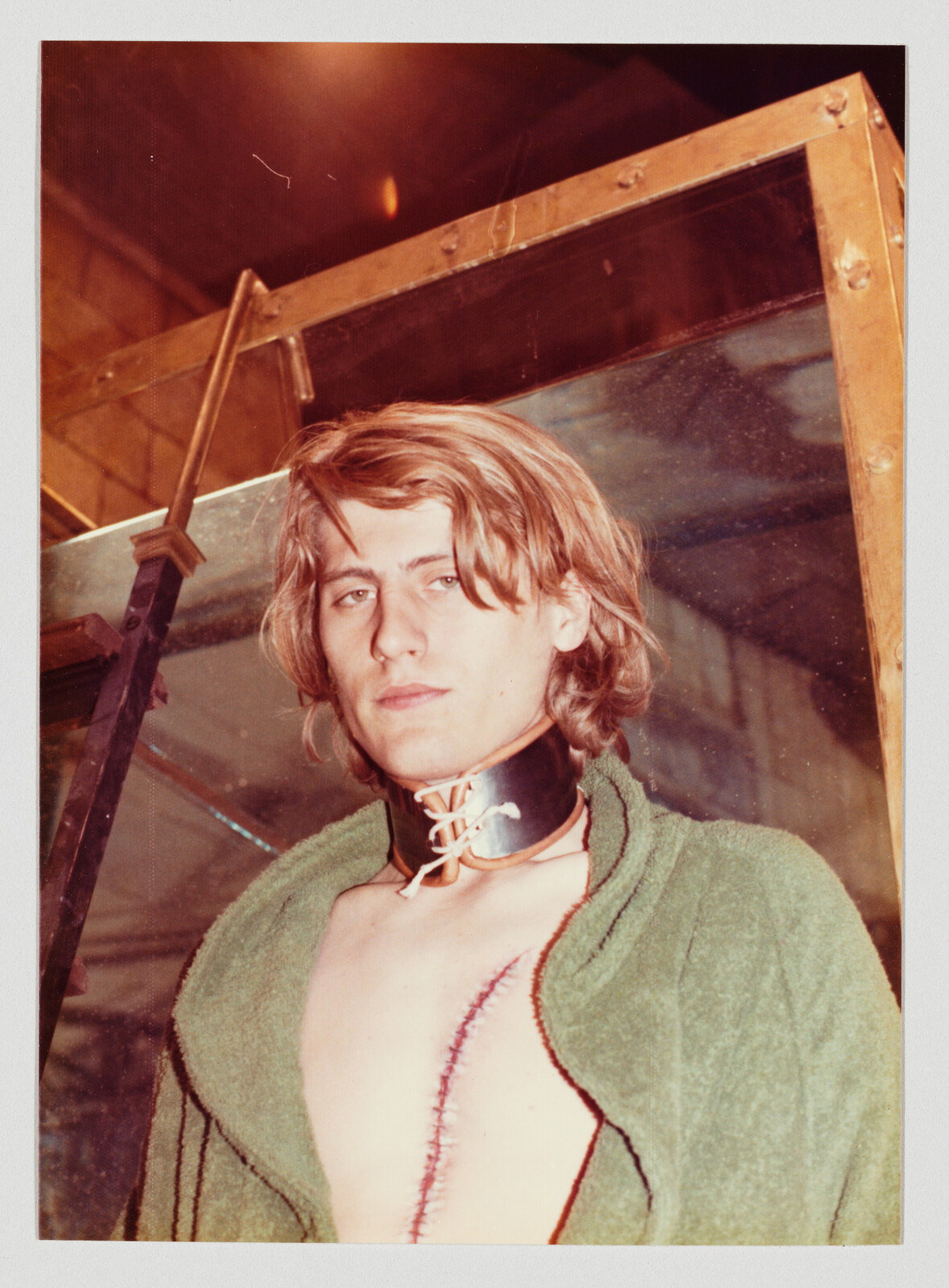 Young man with leather neck brace and large stitched surgical scar down his chest, wearing a green robe.