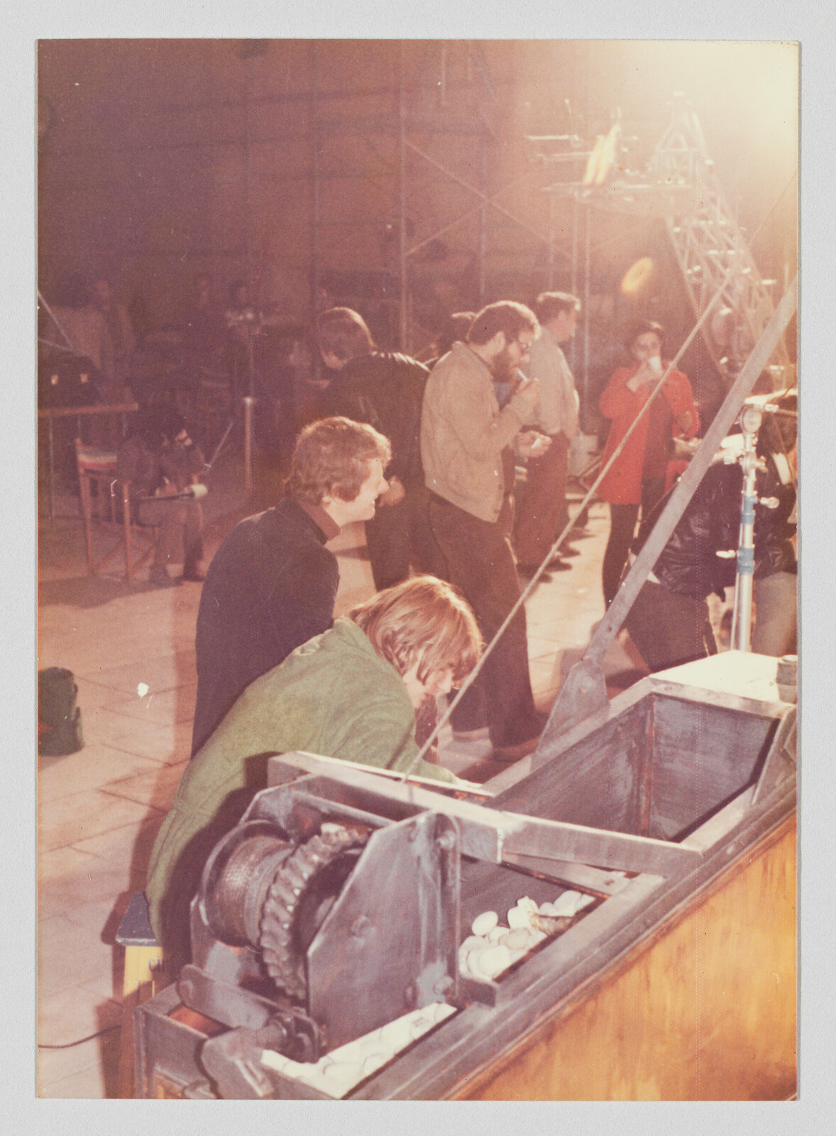 Several crew members inspect a large wooden winch and mechanism while others stand and talk nearby.