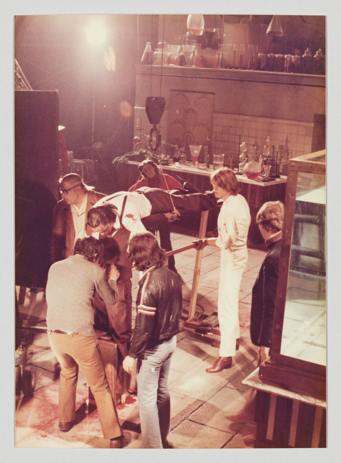 Several people gather around a person lying on a table in a laboratory set, adjusting equipment and props.