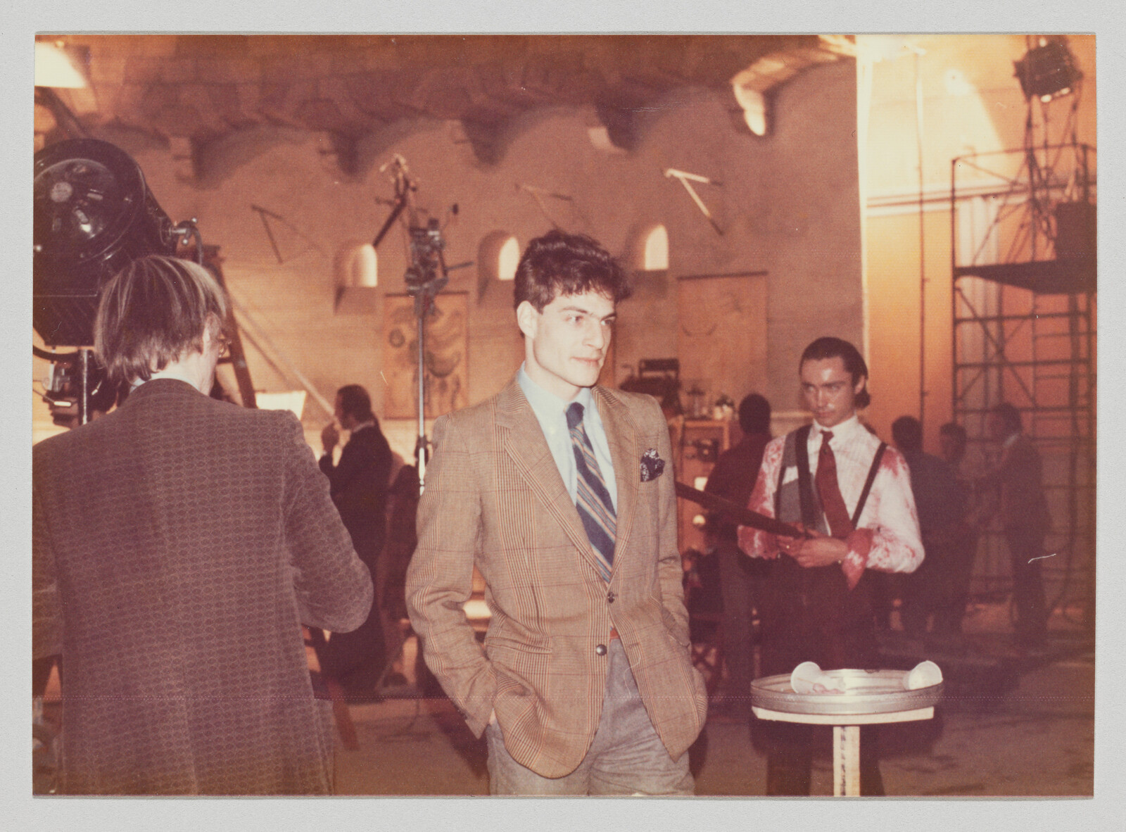 A man in a suit stands with hands in pockets while another man behind him holds a prop weapon.