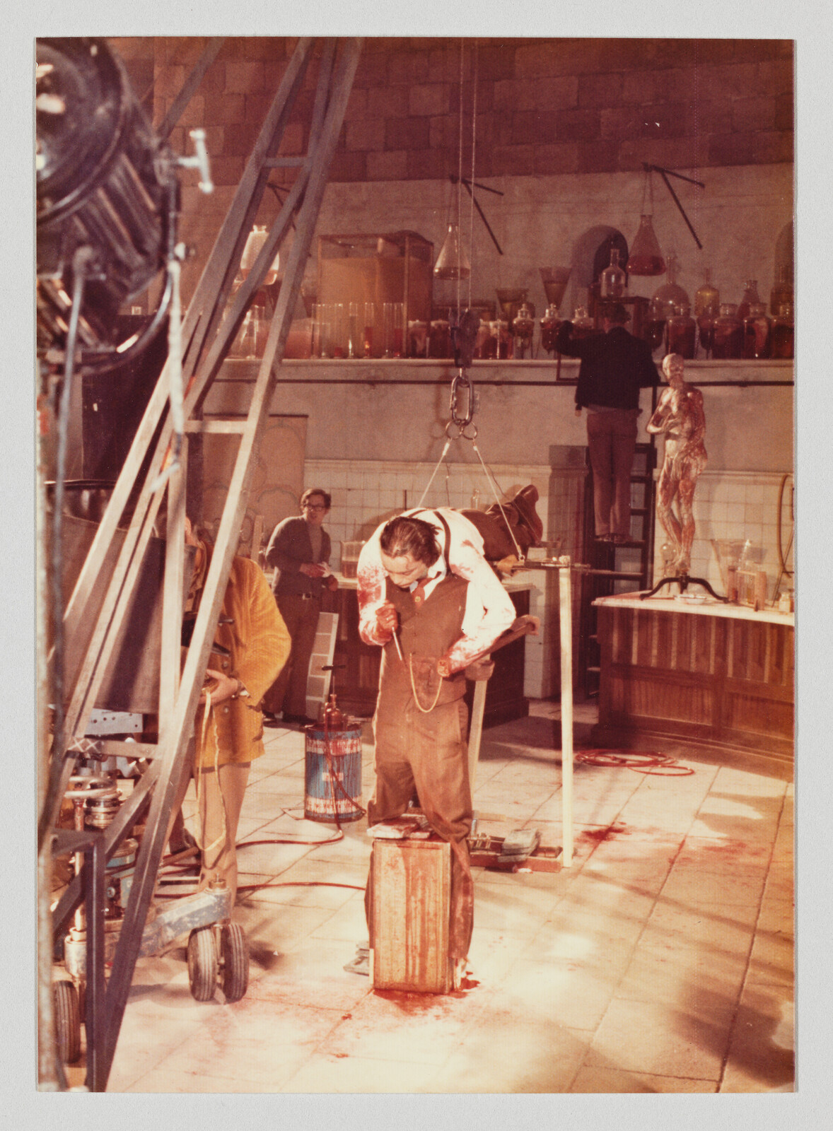 Man in a stained apron leans over a bloodied wooden box inside a laboratory set while crew members work.