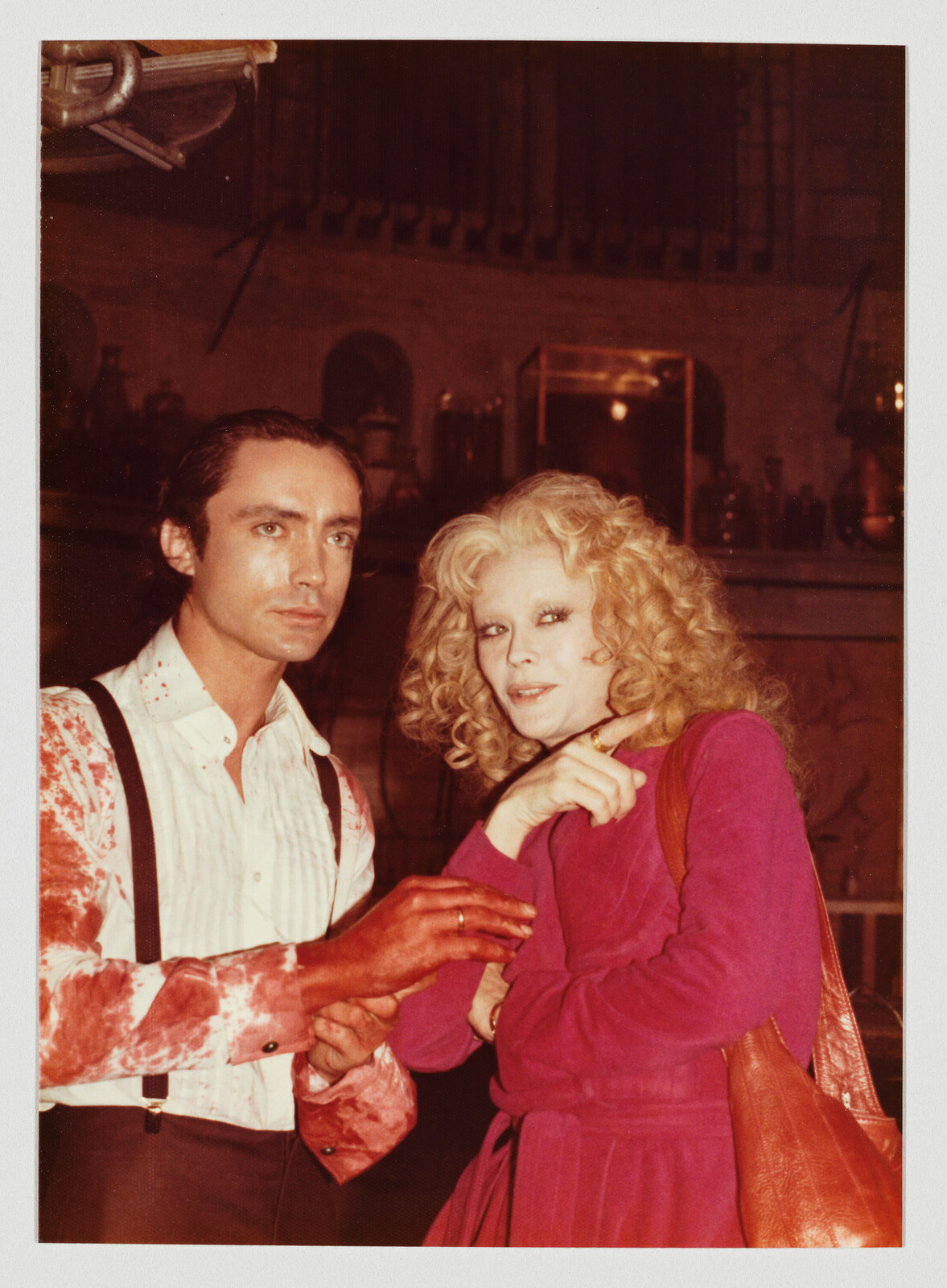 A man with blood-stained shirt and suspenders stands beside a curly-haired woman in a pink robe.