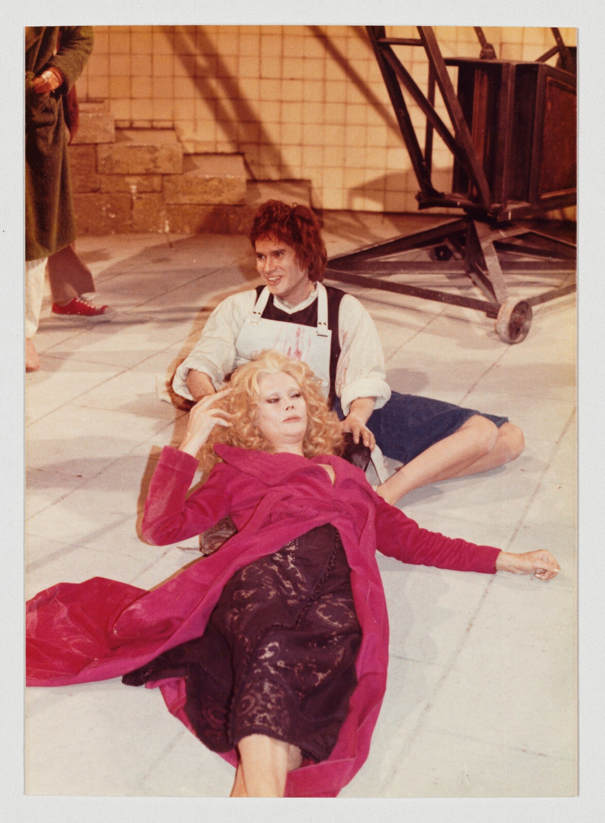 A woman in a bright pink coat lies on the floor while a man behind her gently holds her head.