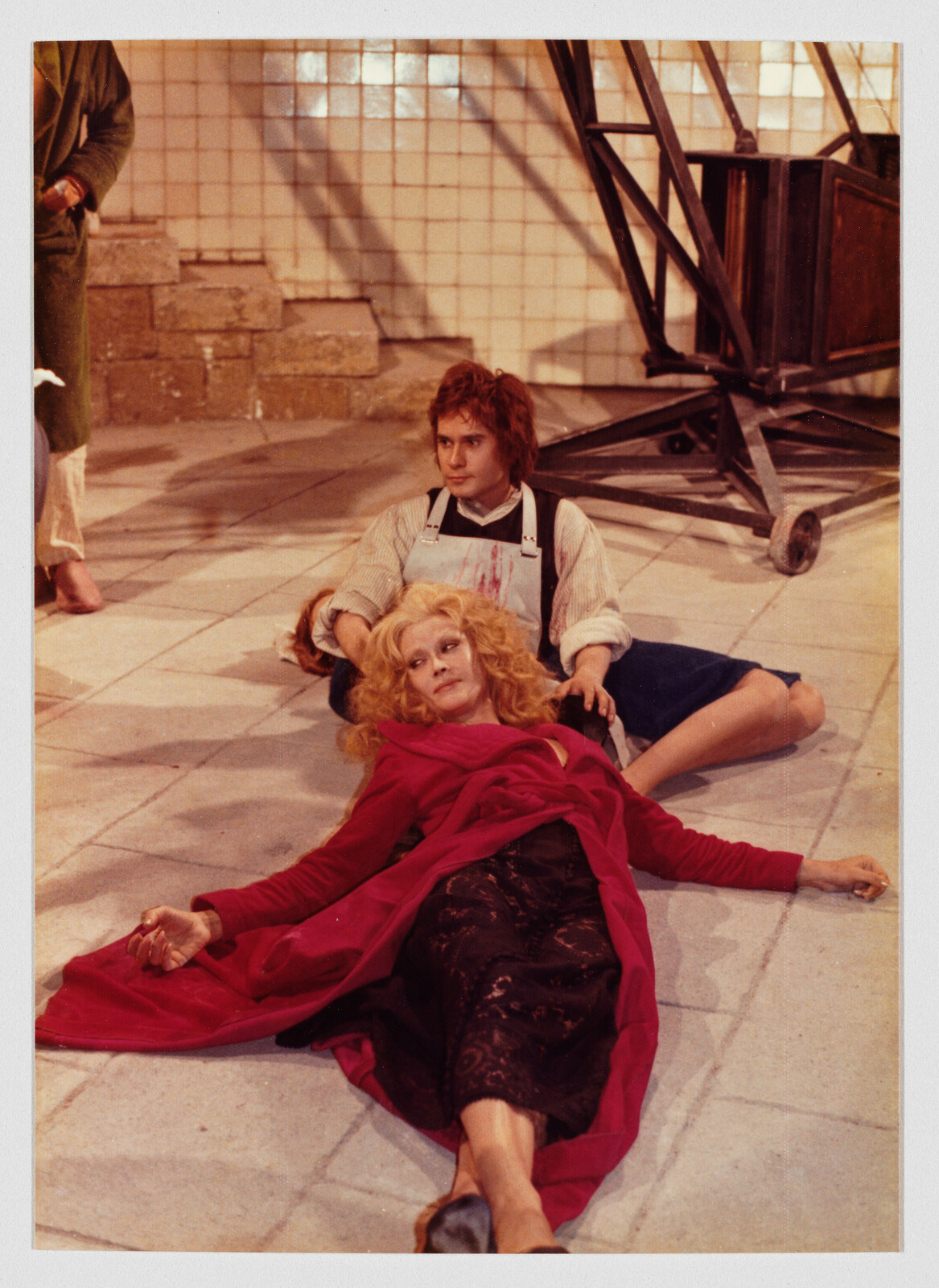 A woman in a red coat lies on the floor while a young man supports her from behind.