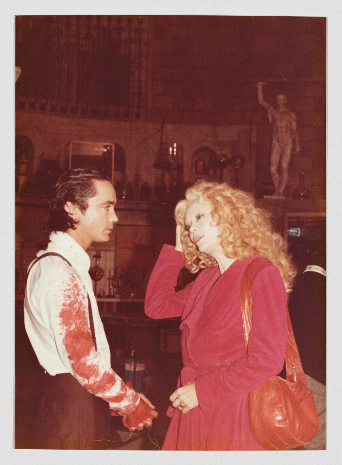 Man with blood-stained sleeve and woman in pink coat talk while she touches her hair.