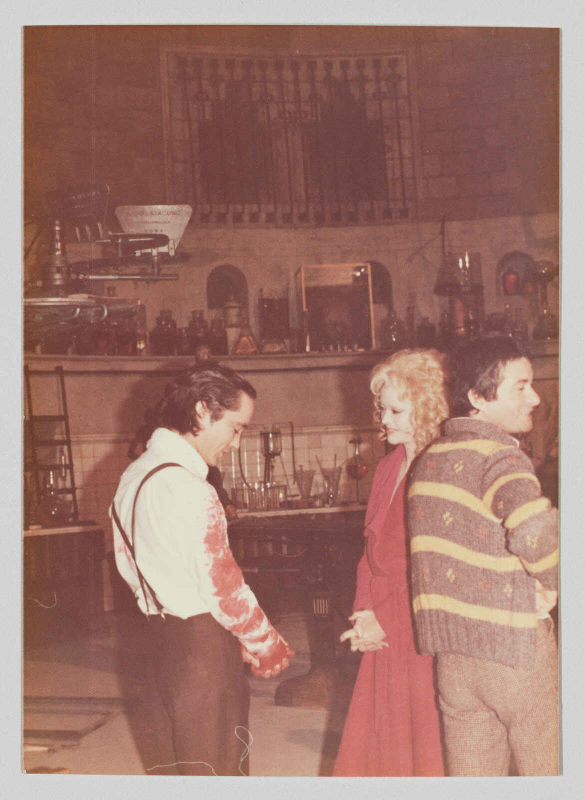 Three people stand in a cluttered laboratory set while one man looks at his blood-stained sleeve.