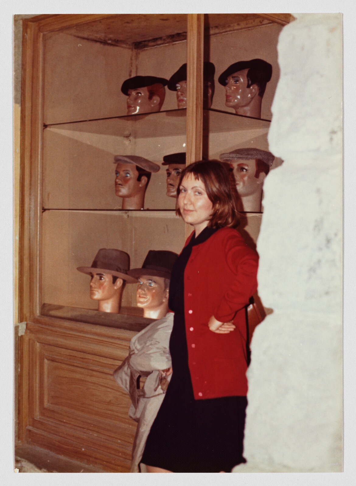 A woman in a red cardigan stands beside a shop display of mannequin heads wearing hats.
