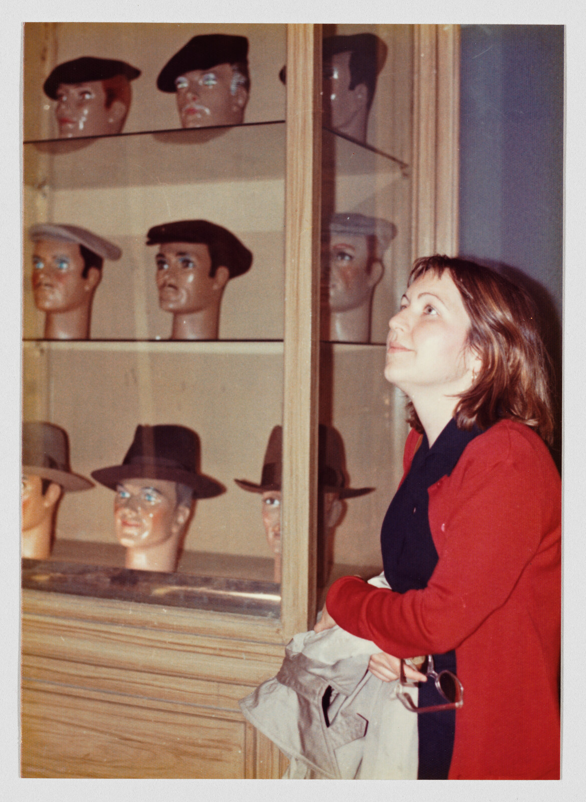 A woman looks up at a glass display of mannequin heads wearing various hats.