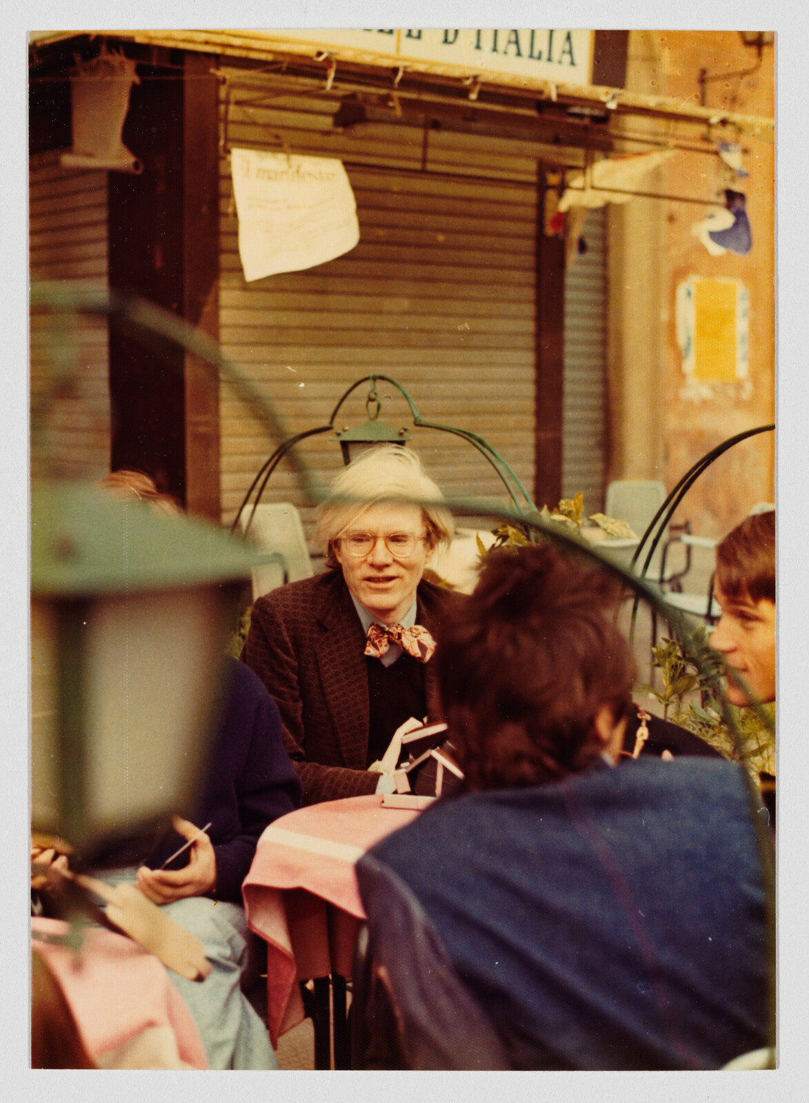 A person with light hair and glasses sits at an outdoor café table talking with others.