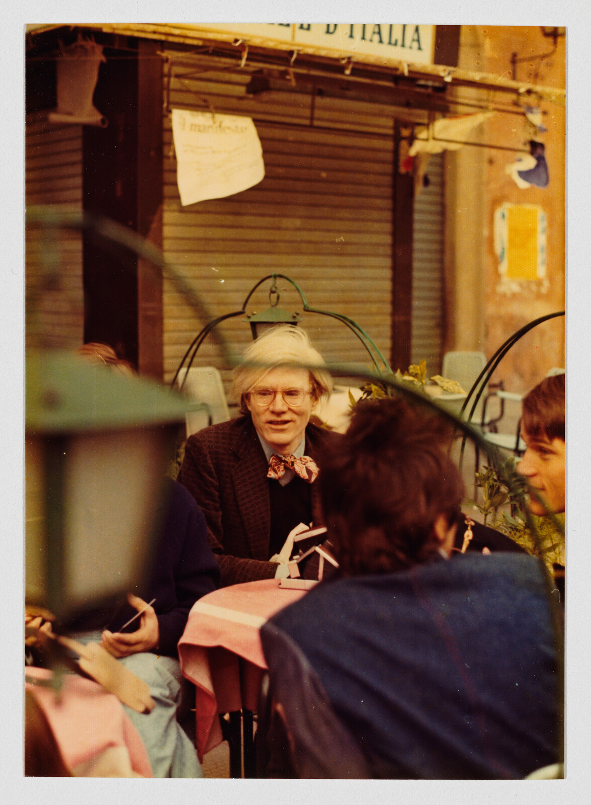 A light-haired person with glasses and a bow tie sitting at an outdoor café table with others.