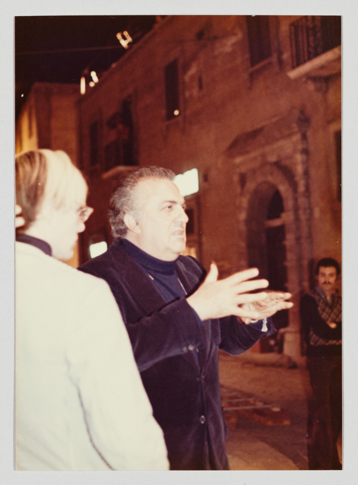 Older man gestures animatedly with both hands while speaking to another person on a dim street.