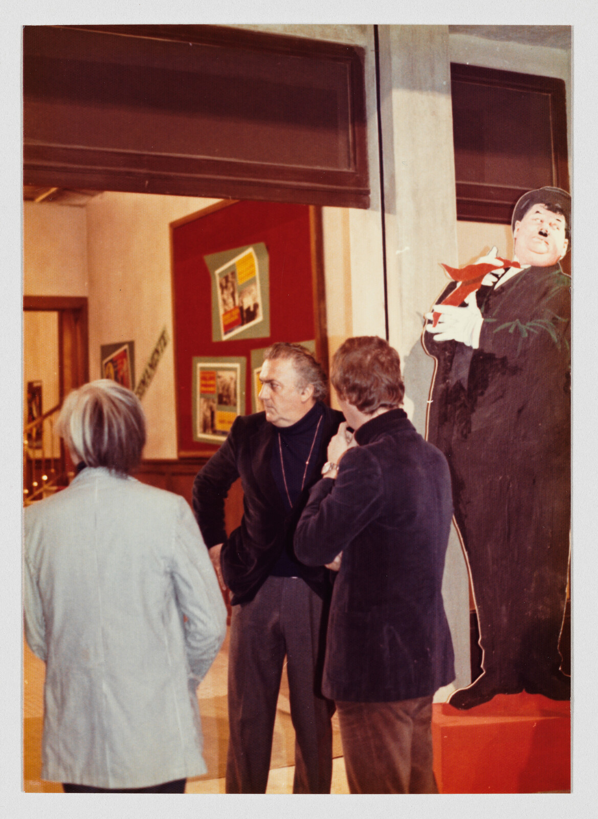 Three men stand in a gallery talking near a large Charlie Chaplin cutout.