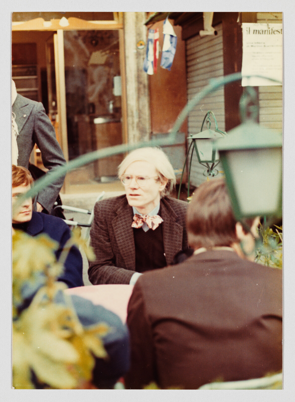 Blond person wearing glasses and a bow tie talks with others at an outdoor café table.