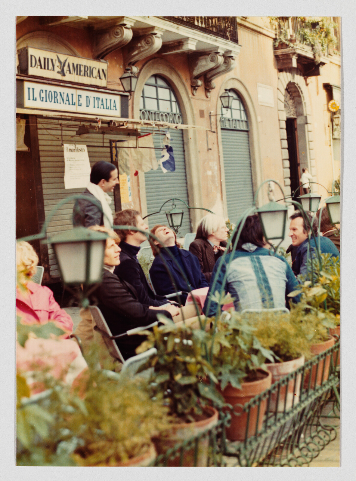 A group of people sit and laugh at an outdoor café in front of closed Italian shopfronts.