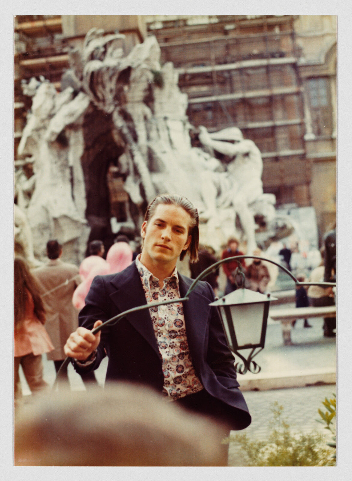 Young man in patterned shirt and blazer holding a curved iron lantern frame in front of a fountain.