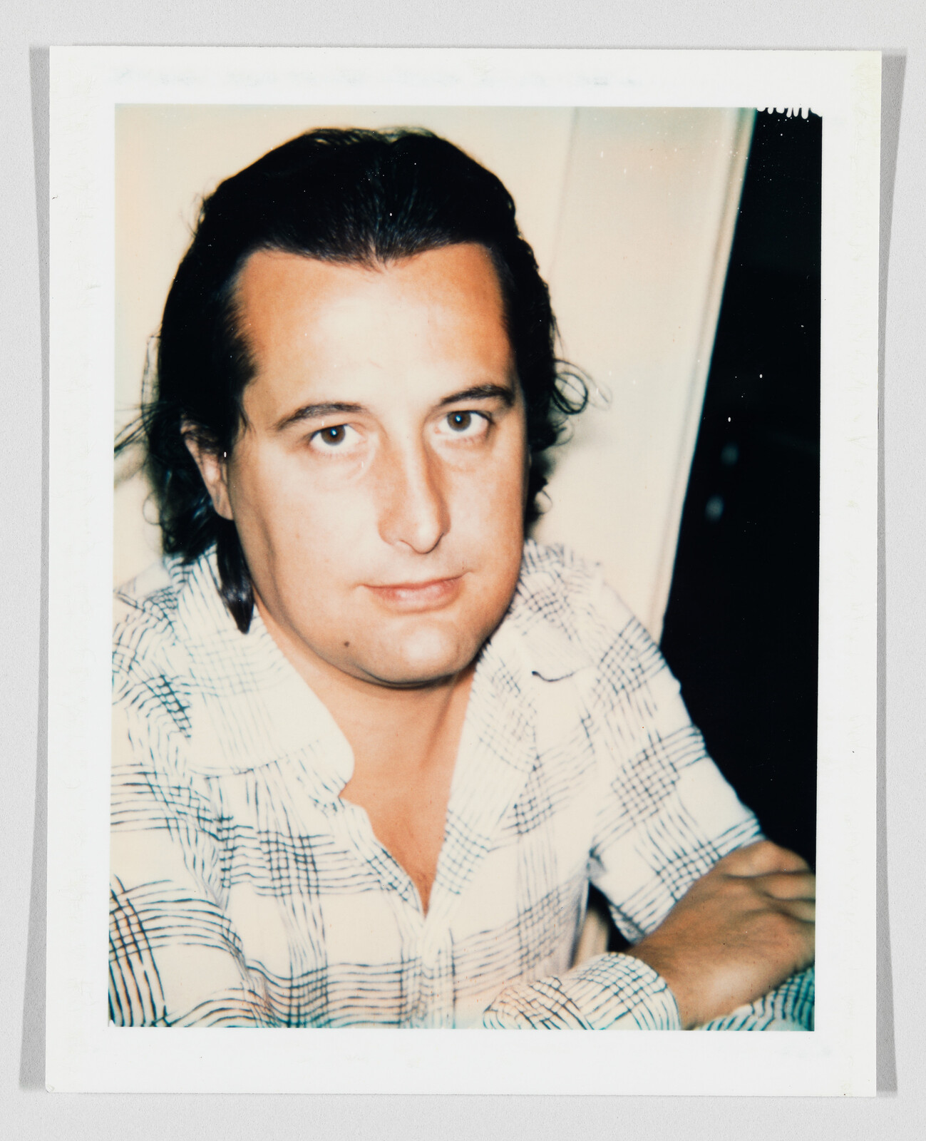 A man with dark hair and a plaid shirt looks at the camera with arms crossed.