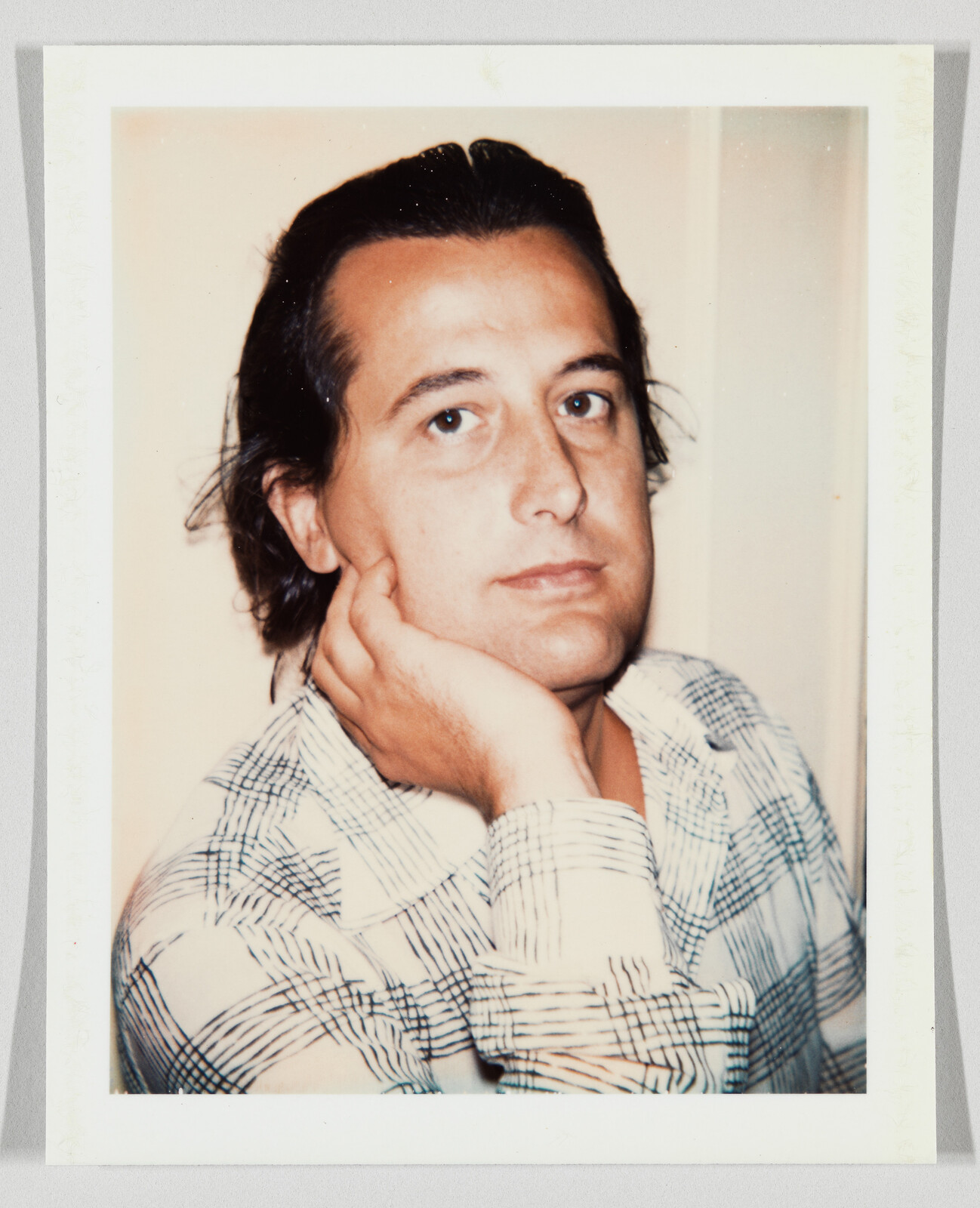 A man rests his cheek on his hand while wearing a patterned button-up shirt.