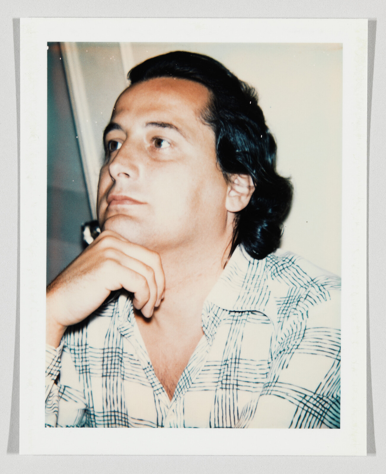 Man with dark hair wearing a patterned shirt rests his chin on his hand, looking thoughtful.