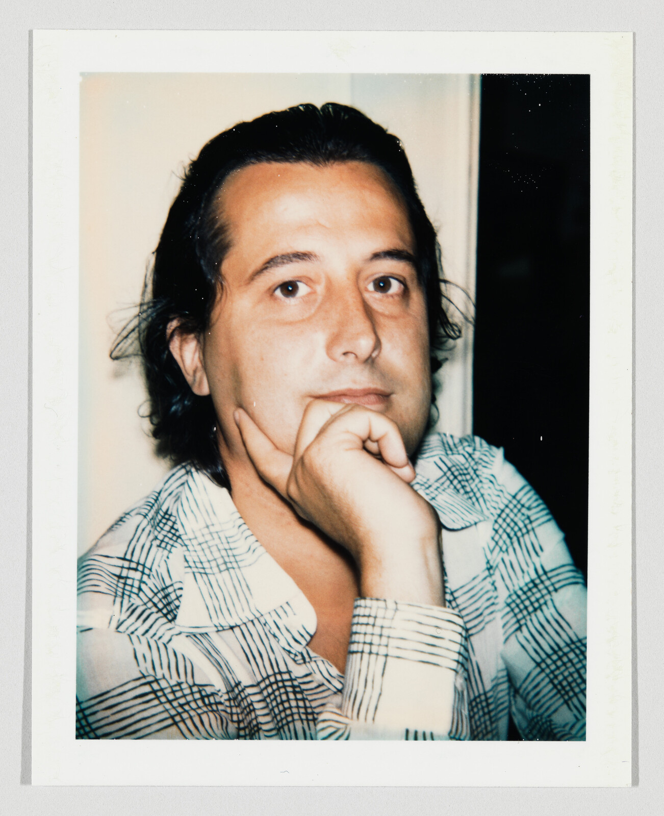 A person with a patterned shirt rests their chin on their hand while looking at the camera.