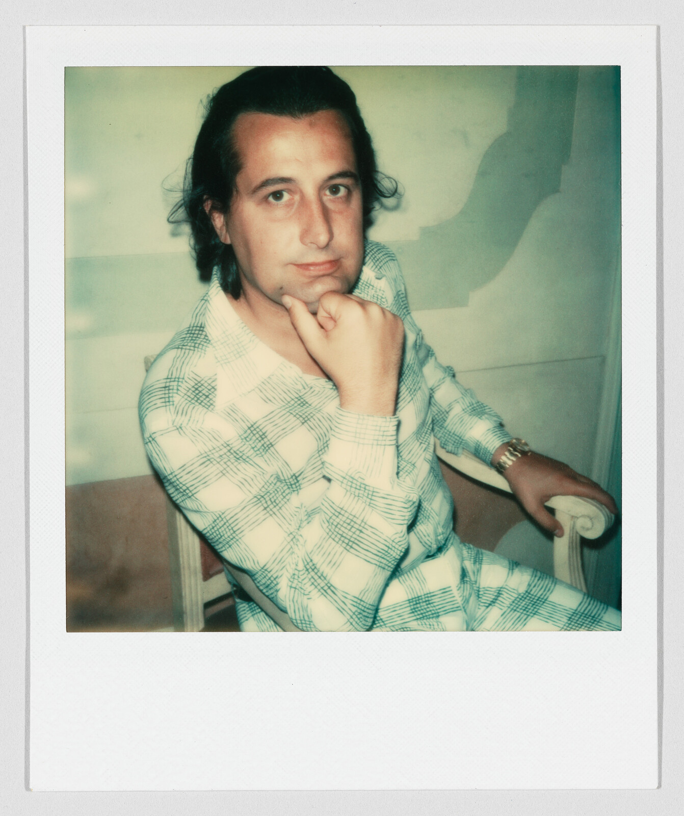 A person with long hair sits on a chair, resting their chin on their hand and looking forward.