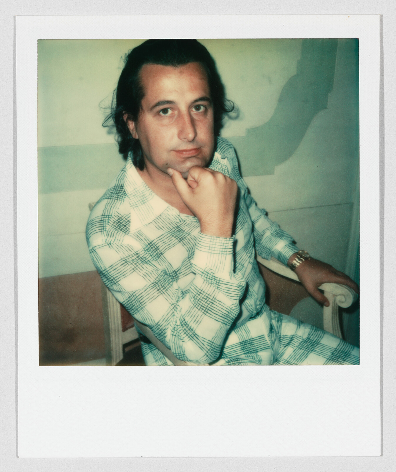 A man in a plaid shirt sits in a chair with his chin resting on his fist.