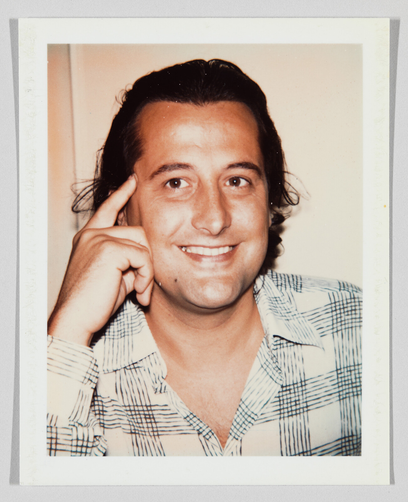 A smiling man touches his temple with one finger while wearing a patterned shirt.
