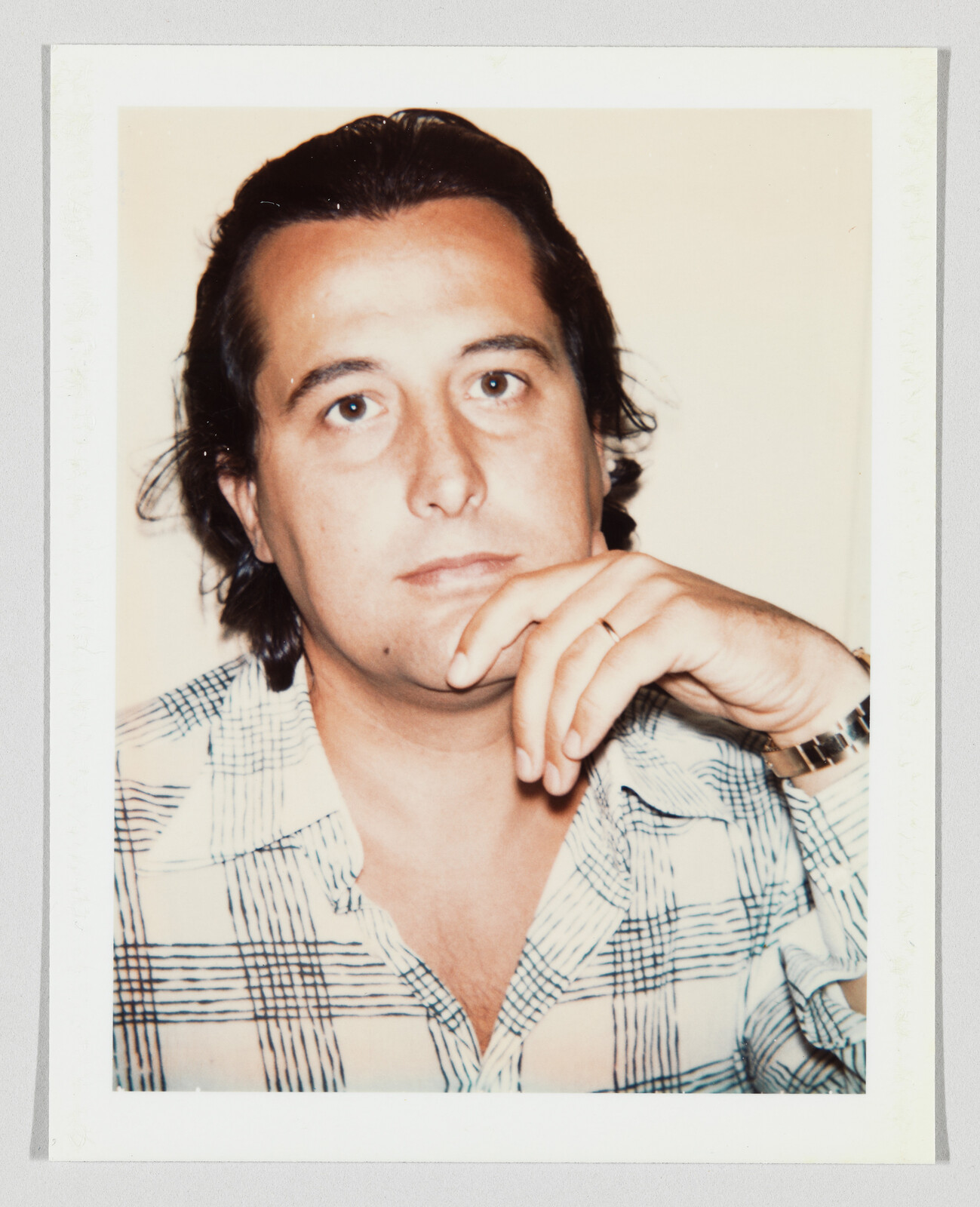 Person with hand on chin wearing a plaid shirt and watch, looking directly at the camera.