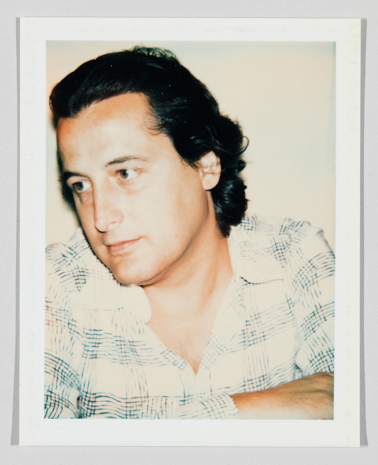 A man with dark hair looks thoughtfully to the left while wearing a checked shirt.