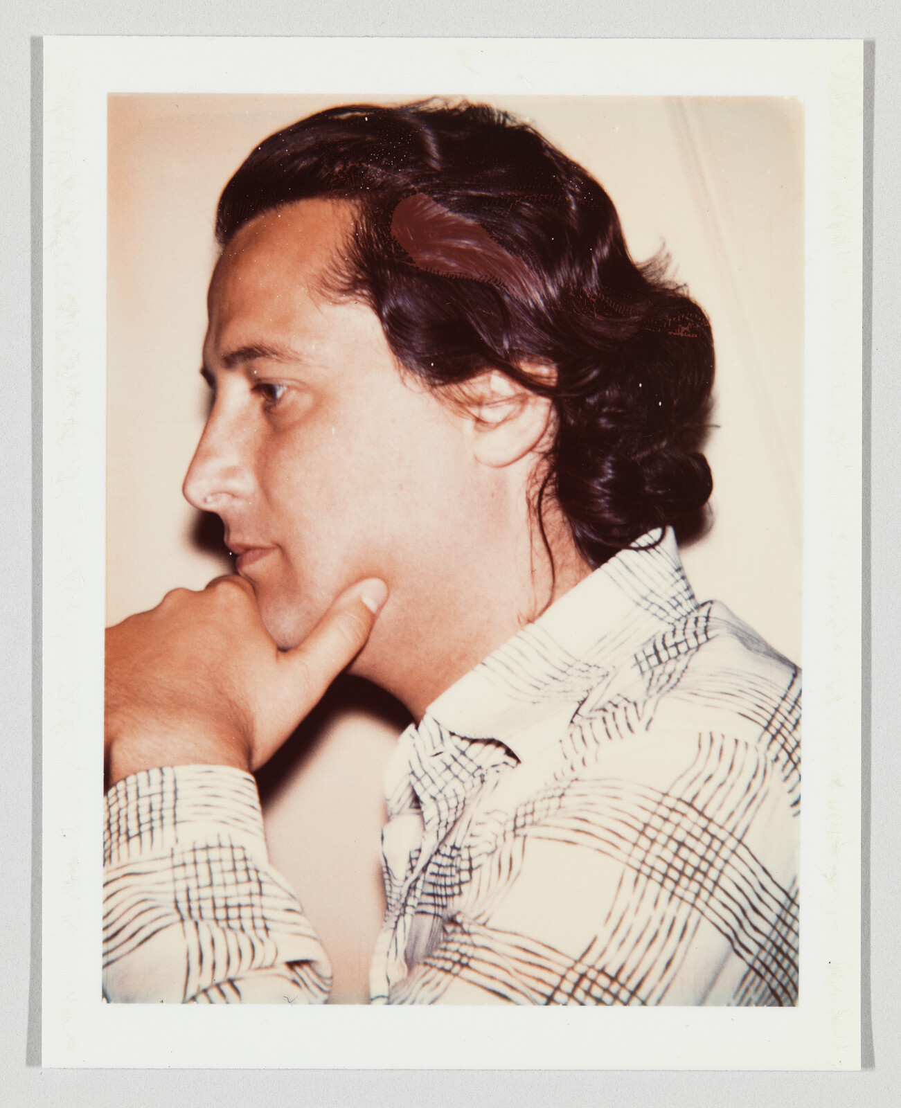 Man with medium-length dark hair rests his chin on his hand, wearing a plaid shirt, looking thoughtful.