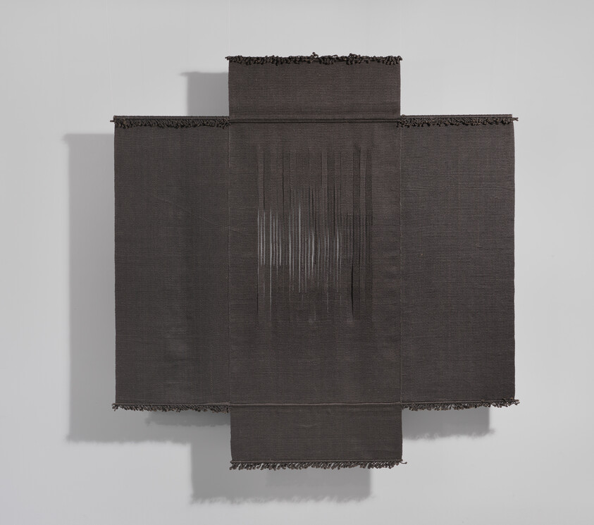A textured black fabric artwork with frayed edges and a vertical white thread pattern on a gallery wall.