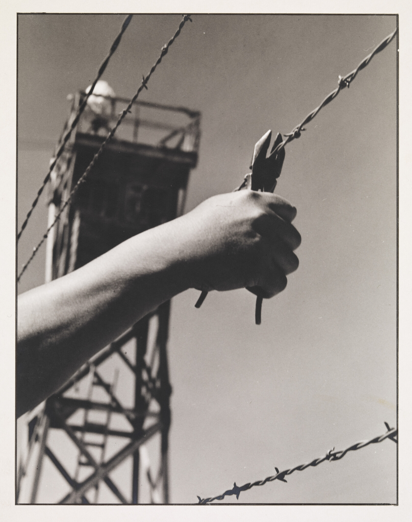 Hand holding wire cutters near barbed wire, with a watchtower in the background.