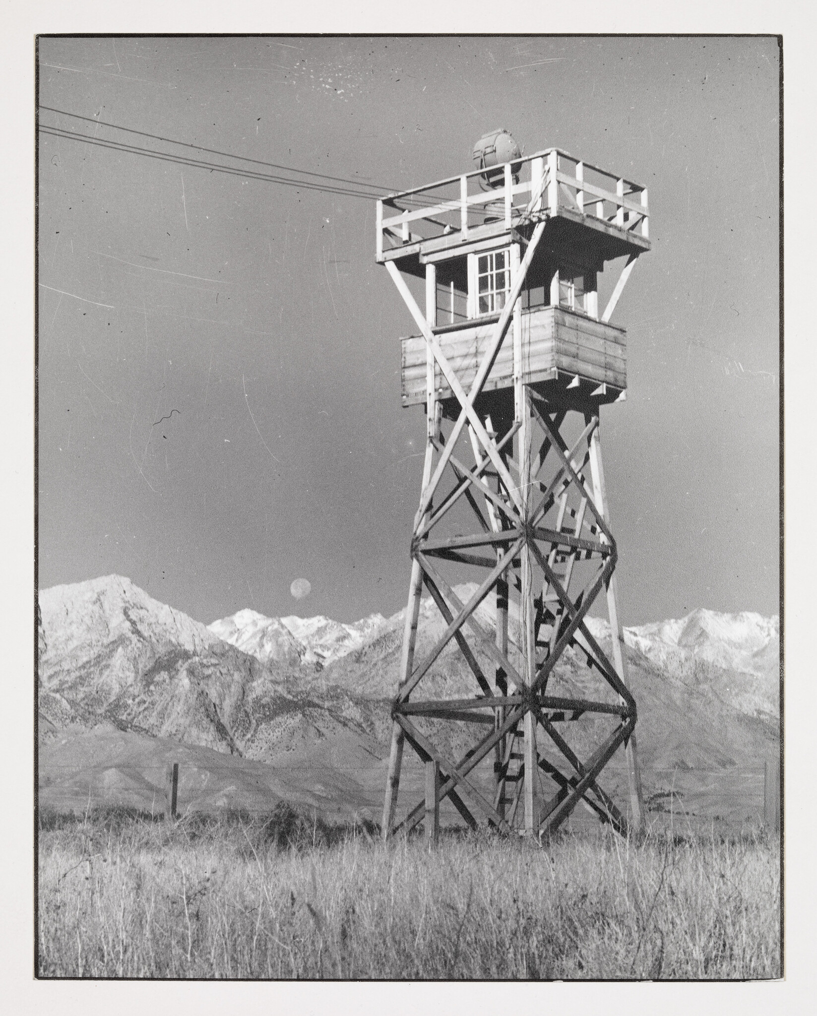 A black and white photograph of a wooden guard tower with a spherical structure on top, standing in a field with a backdrop of rugged mountains and a clear sky. A full moon is visible in the sky above the mountains. The image shows signs of wear with scratches and dust marks.
