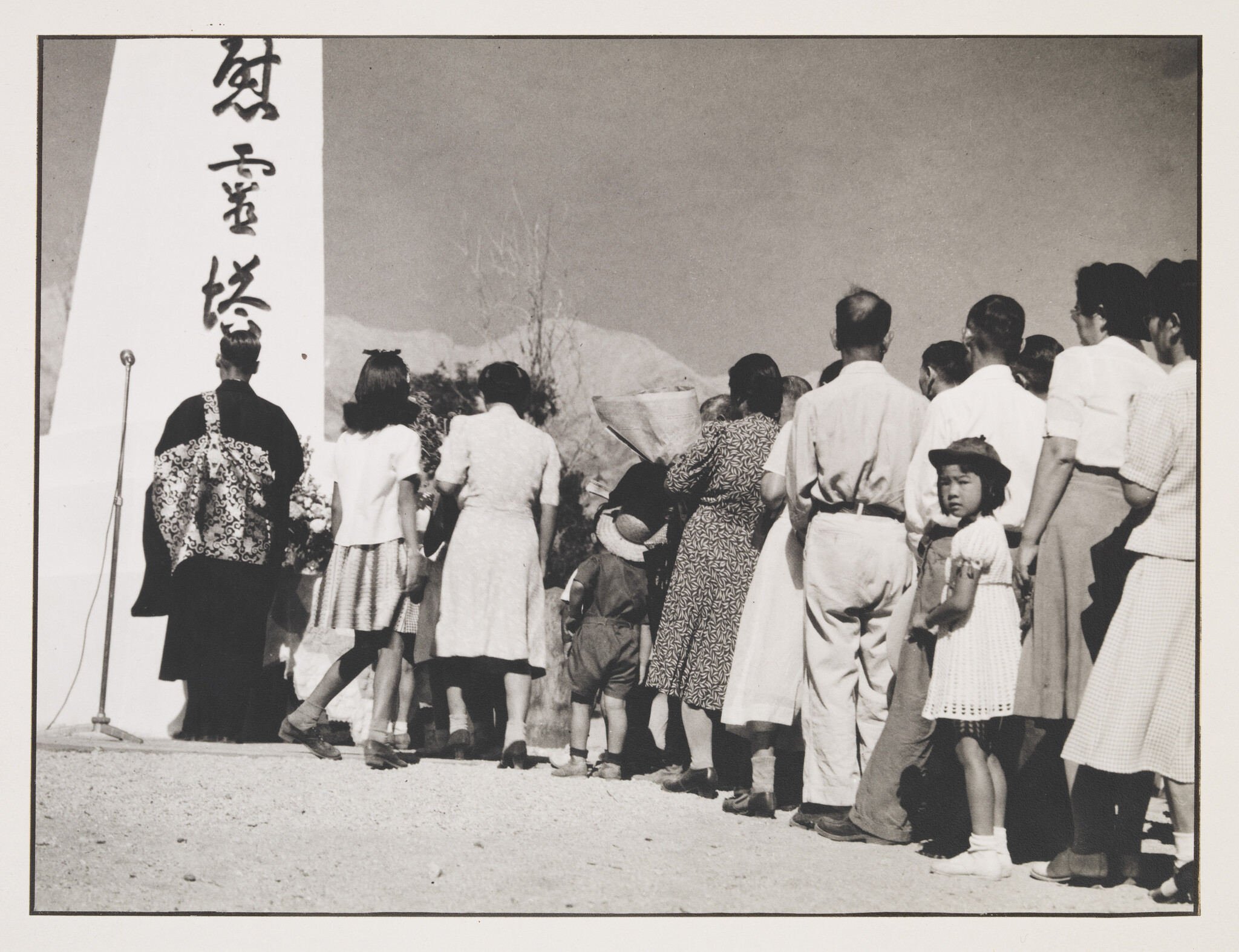 A black and white photograph of a group of people, including children and adults, gathered around a microphone stand next to a tall monument with Japanese characters. The people are dressed in a mix of Western and traditional Japanese attire, suggesting a cultural or commemorative event. A young girl in the foreground looks back towards the camera. The setting appears to be outdoors with a clear sky and sparse vegetation in the background.