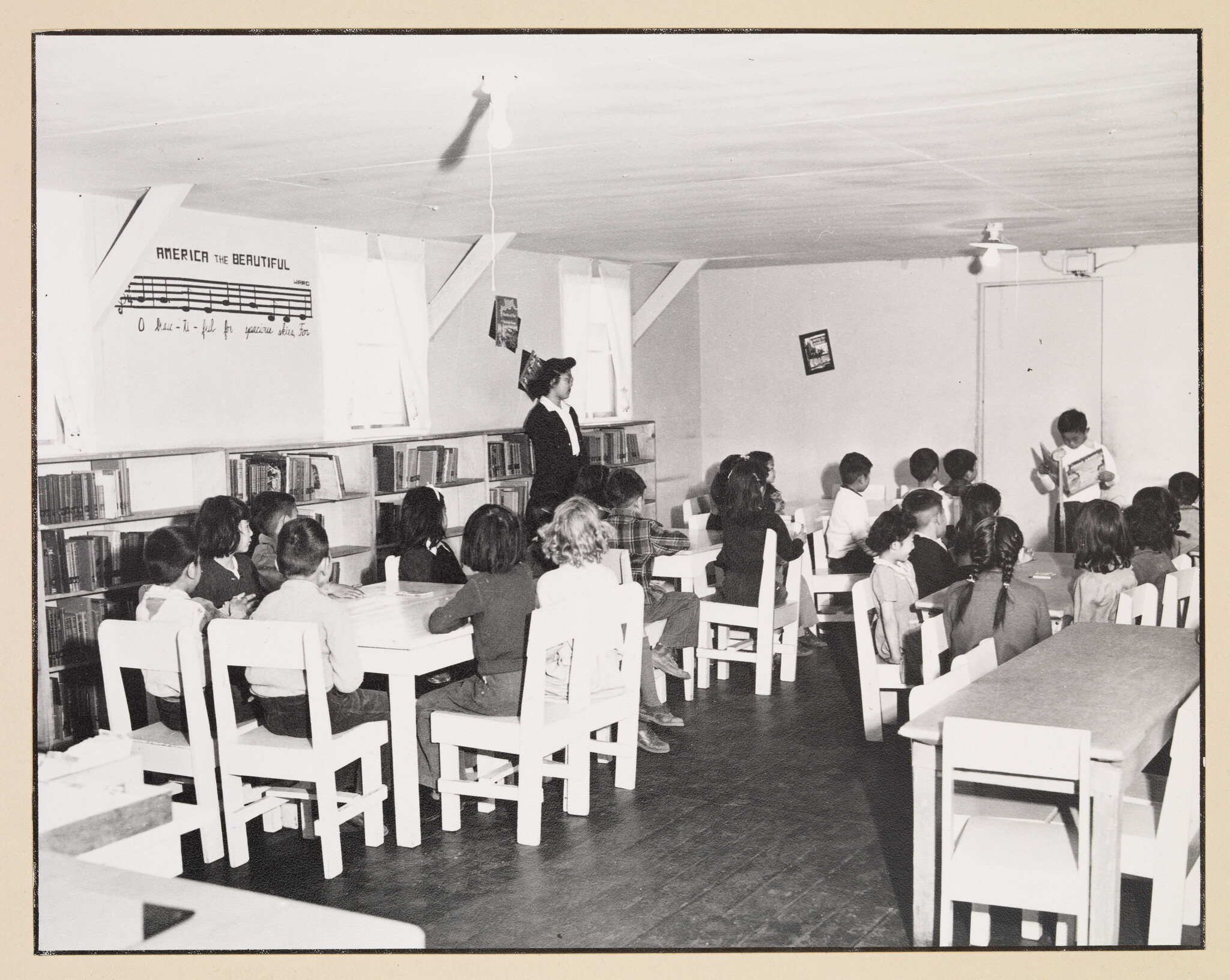 A black and white photo of a classroom scene with students sitting at desks facing a teacher who is standing at the front, reading from a book. The walls are adorned with posters, including one with the musical notes for "America the Beautiful," and bookshelves line the back wall. The setting appears to be from a mid-20th century era.