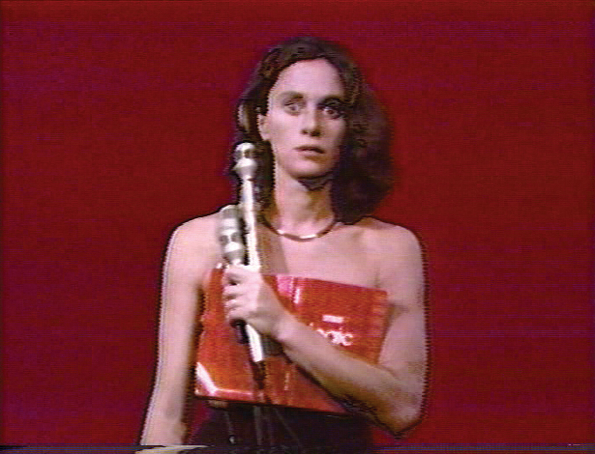 A person wearing a red strapless top holds a microphone and looks straight ahead.