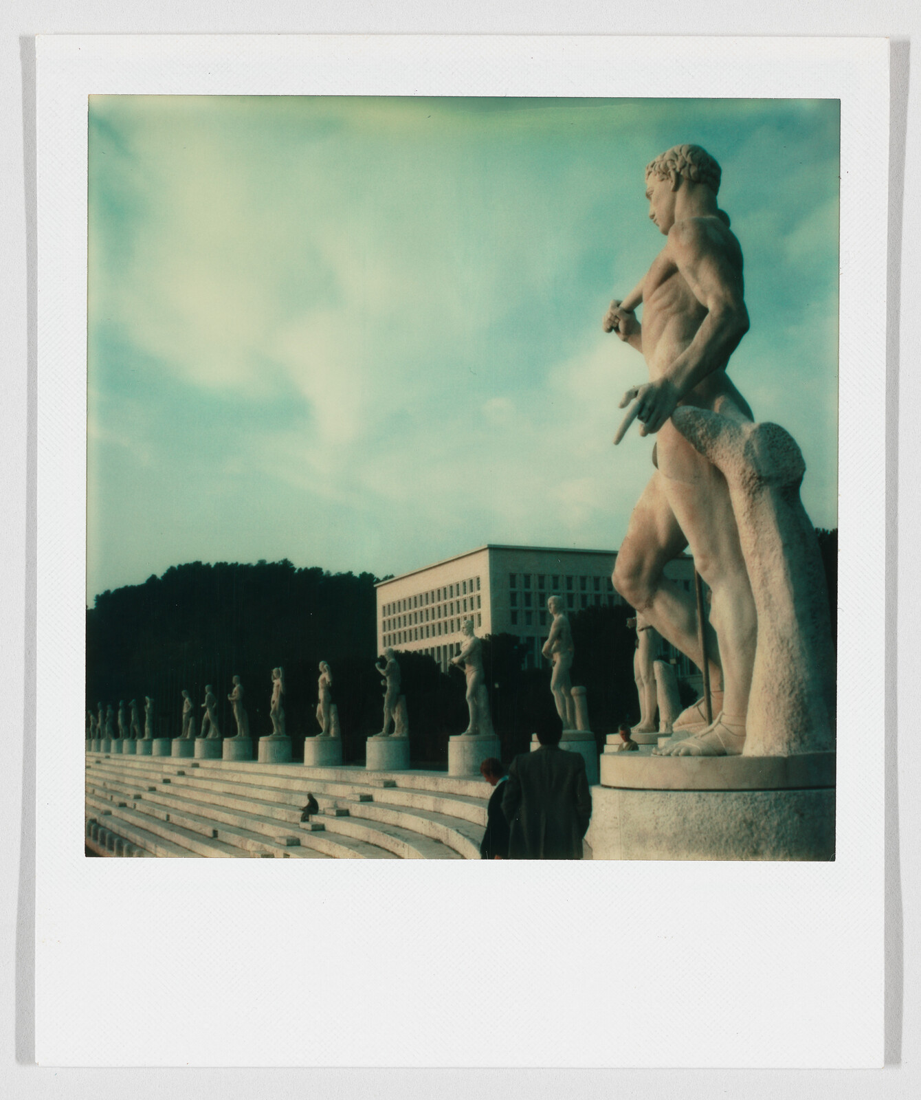 Large stone statue of a nude man stands on steps, with more statues and a modern building in the background.