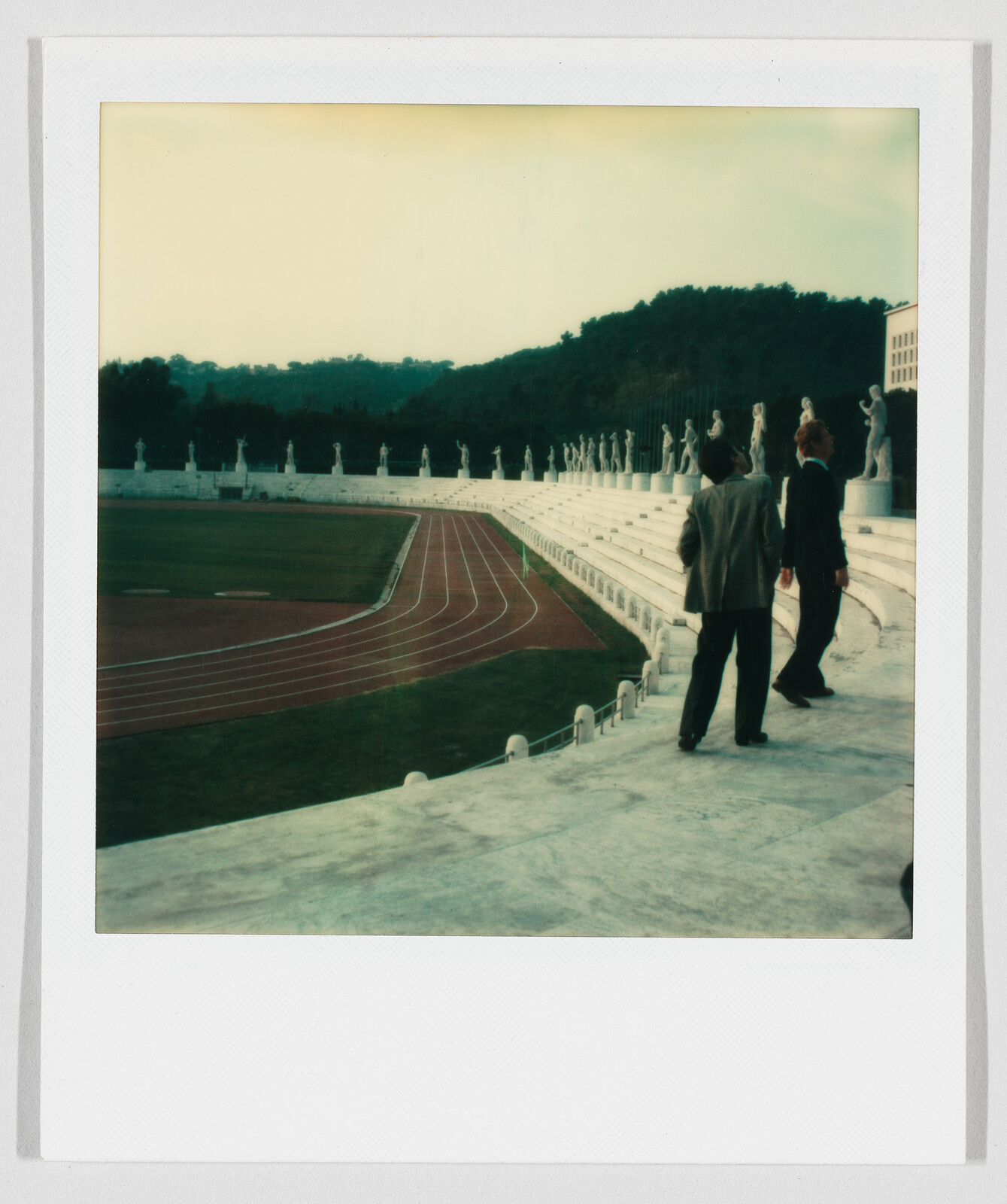 Two people walk along marble stadium steps past a curved running track and row of statues.